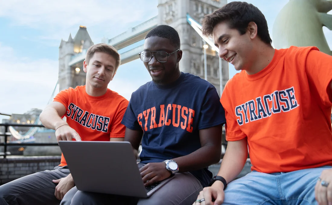 Three students wearing Syracuse University tee shirts sit together in front of the London Bridge.
