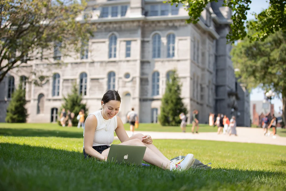 Student sits on grass on laptop.