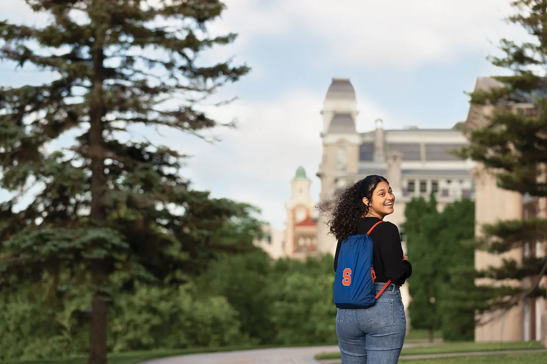 Student wearing a block S logo gray backpack standing on sidewalk in front of Syracuse University Campus buildings crouse college maxell school and hall of languages